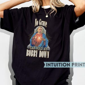 Peut inclure: T-shirt noir avec un graphique d'un homme portant une couronne d'épines tenant un cœur avec le texte "No Grave Can Hold My Bobby Down".
