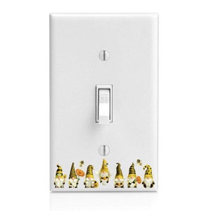 May include: A white light switch cover with a row of seven gnomes wearing yellow and black striped hats. The gnomes are holding flowers and there are bees flying around them.