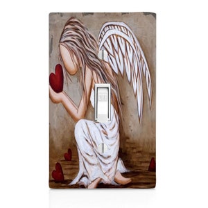 May include: A light switch cover featuring a painting of an angel with wings, kneeling and holding a red heart. The angel is wearing a white dress and has long blonde hair. The background is a brown and beige color with red hearts scattered on the ground.