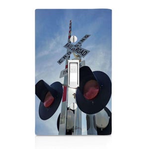 May include: A light switch cover featuring a photograph of a railroad crossing with a blue sky background. The image includes a crossing sign, red signal lights, and the word "OFF" on the switch.