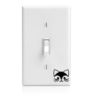 May include: White light switch with a black and white cat face sticker. The sticker has a simple design with a smiling cat face.