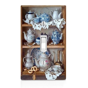 May include: A light switch cover featuring a vintage still life of blue and white teapots, cups, and other porcelain items on a wooden shelf. The image is painted in a realistic style and includes details like lace doilies and pearl necklaces.