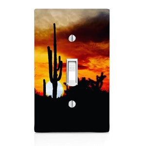 May include: A light switch cover with a sunset over a desert landscape featuring a silhouette of a cactus and other desert plants. The sky is a vibrant orange and yellow with clouds. The switch is labeled "OFF".