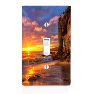 May include: A decorative light switch cover featuring a sunset beach scene with a castle on a cliff. The switch is in the "OFF" position. The sky is a mix of orange, yellow, and blue.