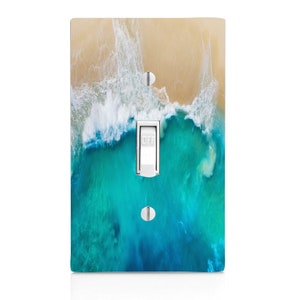 May include: A decorative light switch cover featuring a beach scene with turquoise water and a sandy shore. The switch is white and reads "OFF". The cover is rectangular and has two silver screws.