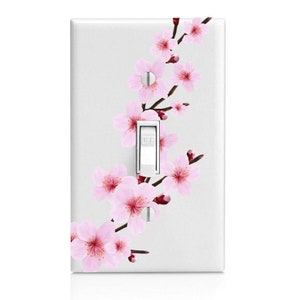 May include: White light switch cover with a pink cherry blossom design. The design features a branch of cherry blossoms with pink flowers and green leaves.