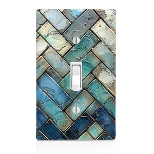 May include: A rectangular light switch plate featuring a herringbone pattern of textured, painted rectangular tiles in shades of blue, teal, and beige. The plate has a central white toggle switch labeled OFF and two silver mounting screws.