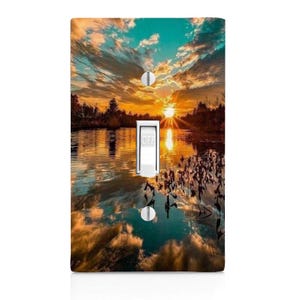 May include: A light switch cover featuring a scenic sunset over a lake with a reflection of the sky and clouds in the water. The sun is setting behind a line of trees on the far shore.
