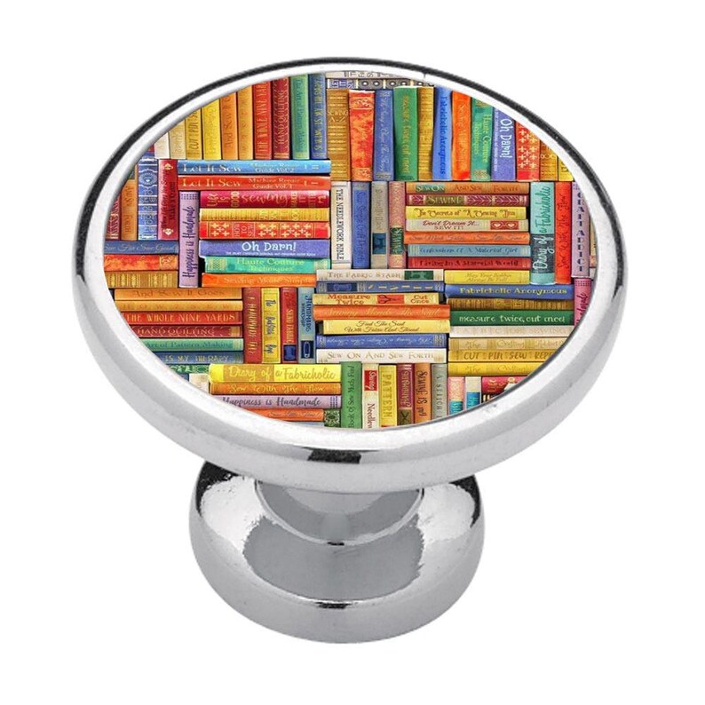 May include: A round, silver-toned metal cabinet knob featuring a flat, circular face decorated with a colourful, dense collage of miniature book spines. The books display various titles and text, including Let It Sew, Oh Darn, and Diary of a Fabricholic.