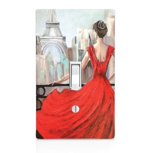 May include: A light switch cover featuring a painting of a woman in a red dress standing on a balcony overlooking the Eiffel Tower in Paris. The painting has a soft, painterly style.