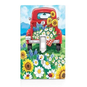 May include: A red vintage pickup truck with a license plate that reads "4T9ER" is filled with flowers. The truck is parked in a field of flowers, including sunflowers, daisies, and bluebonnets. The truck has a red and white checkered picnic blanket in the back. The image is a light switch cover.