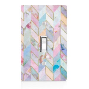 May include: A light switch cover with a pastel watercolor chevron pattern in shades of pink, blue, and lavender, accented with gold lines. The switch is in the "off" position.