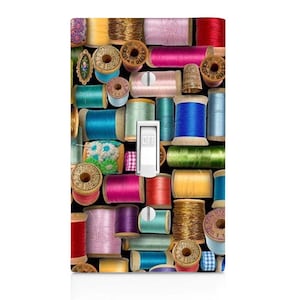 May include: A decorative light switch cover featuring a collage of colorful spools of thread. The spools are in various colors, including pink, blue, green, and gold. The switch is white and in the "off" position.