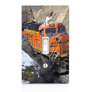 May include: A light switch cover featuring an orange and black BNSF train locomotive. The train is traveling through a mountain pass. The light switch is in the "OFF" position.