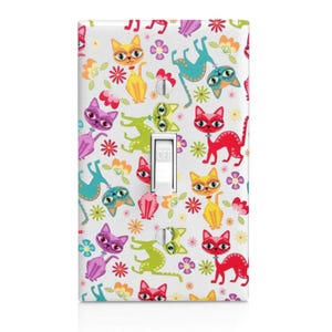 May include: A white light switch cover featuring a colorful pattern of cartoon cats, flowers, and musical notes. The cats are in various colors, including red, yellow, purple, and teal. The switch is in the "off" position.