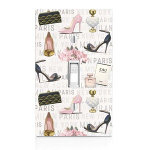 May include: A light switch cover with a white background and a repeating pattern of pink and black high heels, purses, perfume bottles, and flowers. The text "Paris, New York, Milan" is repeated throughout the design.