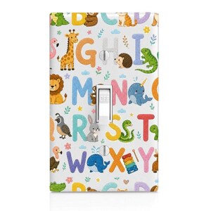May include: A rectangular light switch cover plate featuring a white background with a colorful alphabet pattern. Each letter is paired with a cartoon animal or object, including a giraffe, lion, narwhal, whale, and xylophone, with a central toggle switch.