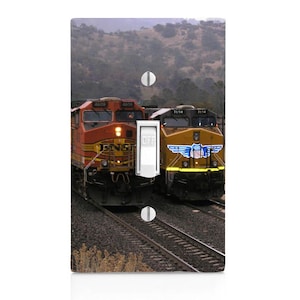 May include: A light switch cover featuring a photograph of two freight trains on a track. The train on the left is red and yellow with the numbers 4605 and 4609 on the side. The train on the right is yellow and brown with the numbers 7614 and 7614 on the side.
