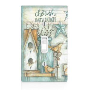May include: A light switch cover with a rustic farmhouse design. The cover features a birdhouse, a vase with flowers, and two birds. The text "cherish every moment" is printed on the cover.