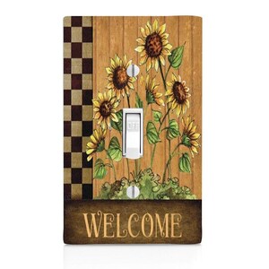 May include: A light switch cover with a woodgrain background and a checkered border. The cover features a watercolor illustration of sunflowers with the word "WELCOME" at the bottom.