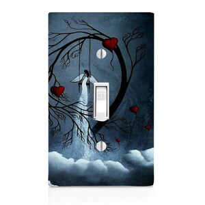 May include: Decorative light switch cover featuring a whimsical design. The artwork depicts an angel on a swing, surrounded by red hearts and a tree silhouette against a dark blue background. The switch is in the "OFF" position.