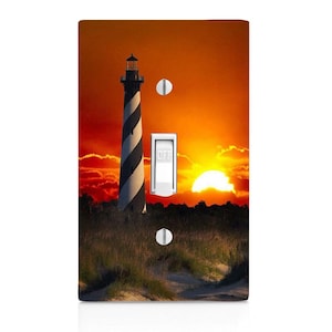 May include: A light switch cover featuring a black and white striped lighthouse silhouetted against a vibrant orange sunset. The lighthouse is on a sandy beach with tall grass.