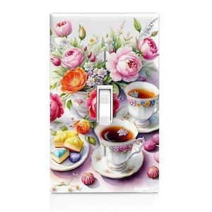 May include: A light switch cover with a floral design featuring pink, orange, and yellow flowers. Two teacups with saucers and a plate of pastries are on a table with a purple tablecloth.