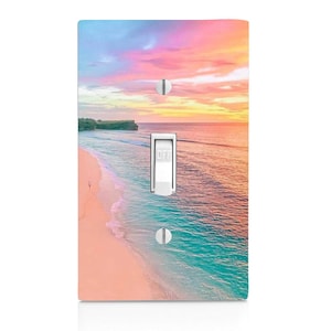 May include: A light switch cover with a colorful sunset over a beach scene. The sky is pink, orange, and blue, and the water is turquoise. A sandy beach is in the foreground.