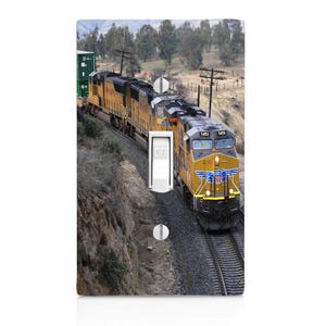 May include: A decorative light switch cover featuring a yellow and black train on a track. The train is traveling through a mountainous landscape with trees and power lines. The switch is in the off position.