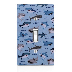 May include: A light blue light switch cover with a pattern of cartoon sharks in various colors and styles. The switch is labeled "OFF".