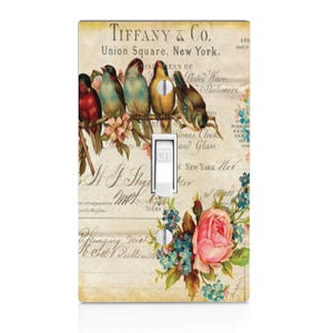 May include: A vintage-style light switch cover featuring a floral design with pink roses and blue flowers. The background is a faded cream color with a handwritten inscription that reads "TIFFANY & CO. Union Square, New York."