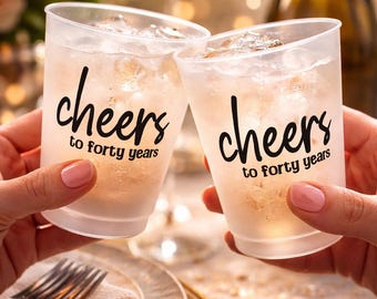 Op de afbeelding: Twee bevroren plastic bekers gevuld met ijs en een lichtgekleurde drank worden omhooggehouden in een feestelijke toast. De bekers zijn bedrukt met de woorden "cheers to forty years" in een zwart scriptlettertype. De achtergrond is zacht vervaagd met warme verlichting.