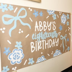 May include: A brown paper banner with the text "ABBY'S eighteenth BIRTHDAY" in white and light blue. The banner is decorated with light blue bows and floral designs. A framed photo collage hangs above.