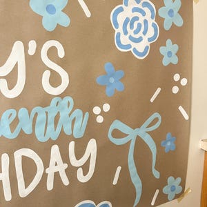 May include: A brown paper poster with the text "'s seventh birthday" in white and light blue. The poster is decorated with light blue flowers, bows, and white dots and dashes. The poster is taped to a wall.