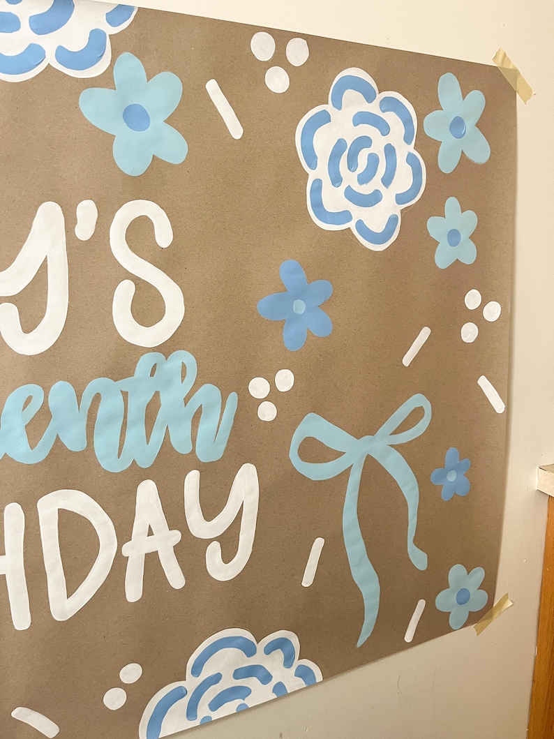 May include: A brown paper poster with the text "'s seventh birthday" in white and light blue. The poster is decorated with light blue flowers, bows, and white dots and dashes. The poster is taped to a wall.