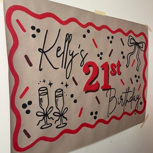 May include: A brown paper banner with red and black accents, celebrating "Kelly's 21st Birthday." The design includes a red border, champagne glasses, a bow, and decorative sprinkles. The banner is taped to a white wall.