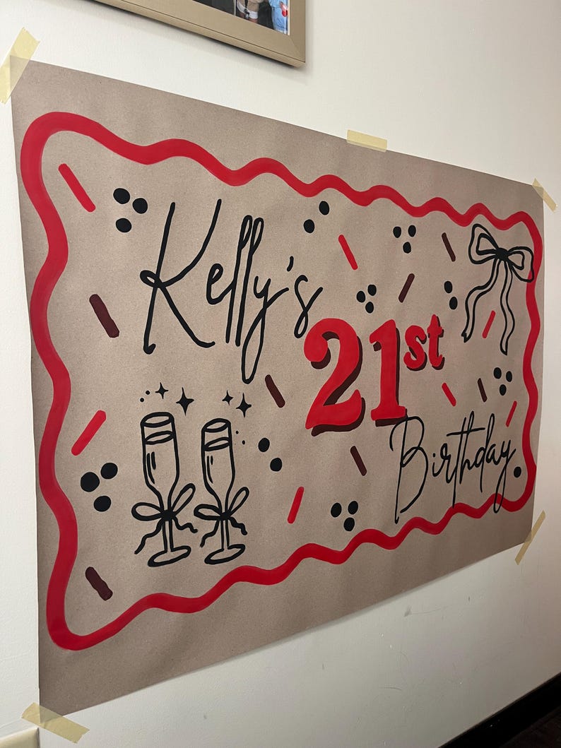 May include: A brown paper banner with red and black accents, celebrating "Kelly's 21st Birthday." The design includes a red border, champagne glasses, a bow, and decorative sprinkles. The banner is taped to a white wall.