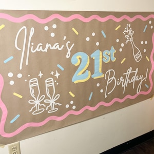 May include: A brown paper banner with the text "Iiana's 21st Birthday!" in white script and blue and yellow block letters. The banner has a pink wavy border and confetti-like decorations, including champagne glasses and a bottle.