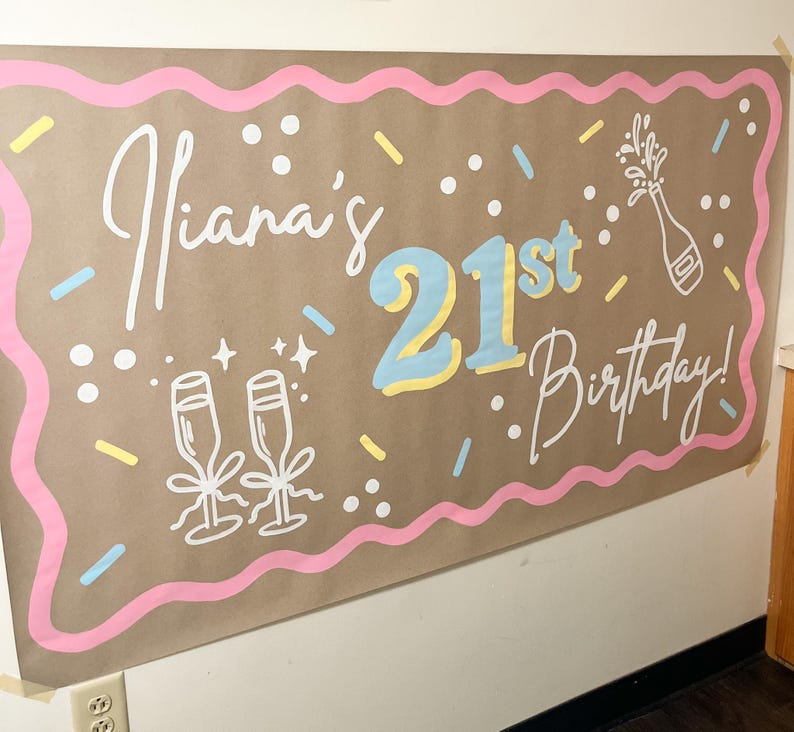 May include: A brown paper banner with the text "Iiana's 21st Birthday!" in white script and blue and yellow block letters. The banner has a pink wavy border and confetti-like decorations, including champagne glasses and a bottle.