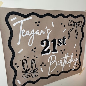 May include: A brown poster with black and white text that reads "Teagan's 21st Birthday." The poster has a black border and includes illustrations of champagne glasses and a bow.