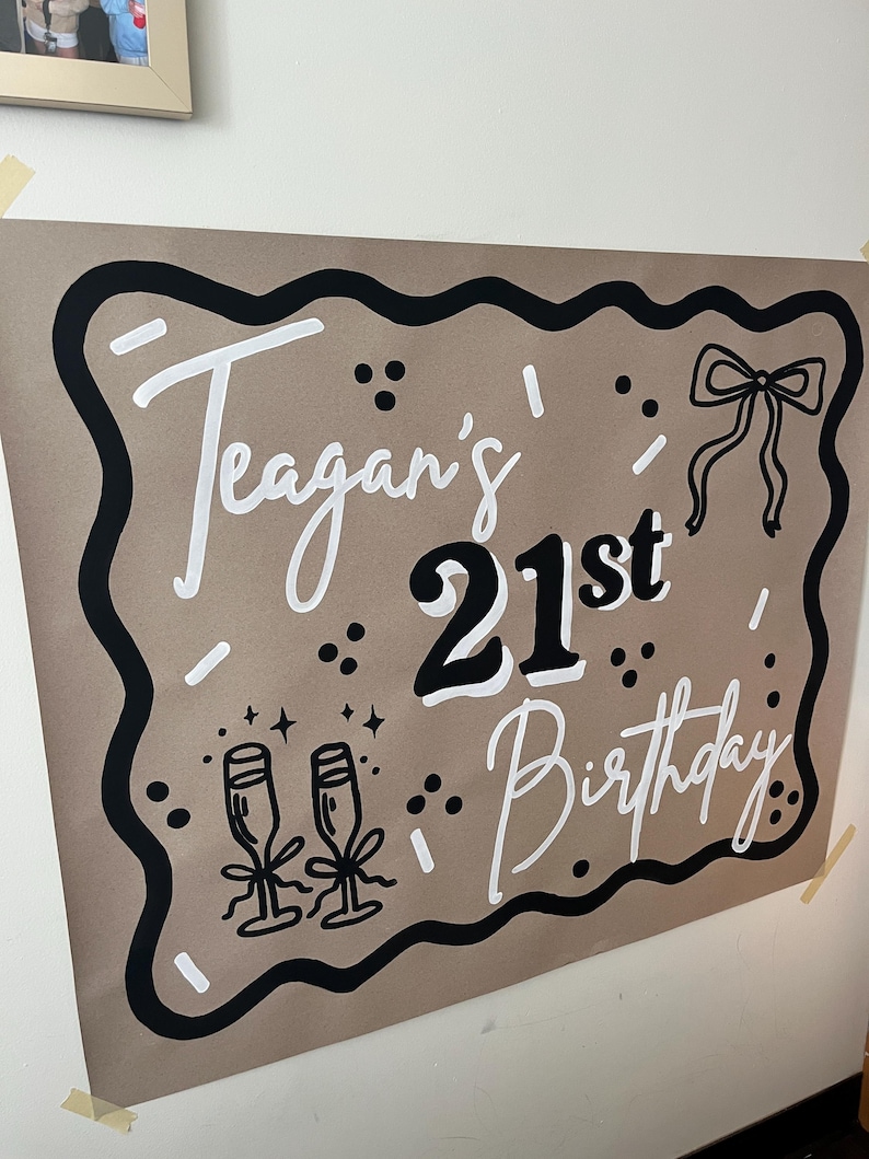 May include: A brown poster with black and white text that reads "Teagan's 21st Birthday." The poster has a black border and includes illustrations of champagne glasses and a bow.