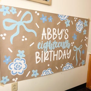 May include: A large brown paper banner with the words "ABBY'S eighteenth BIRTHDAY" in white and light blue lettering. The banner is decorated with light blue bows, flowers, and confetti-like accents. The banner is taped to a white wall.