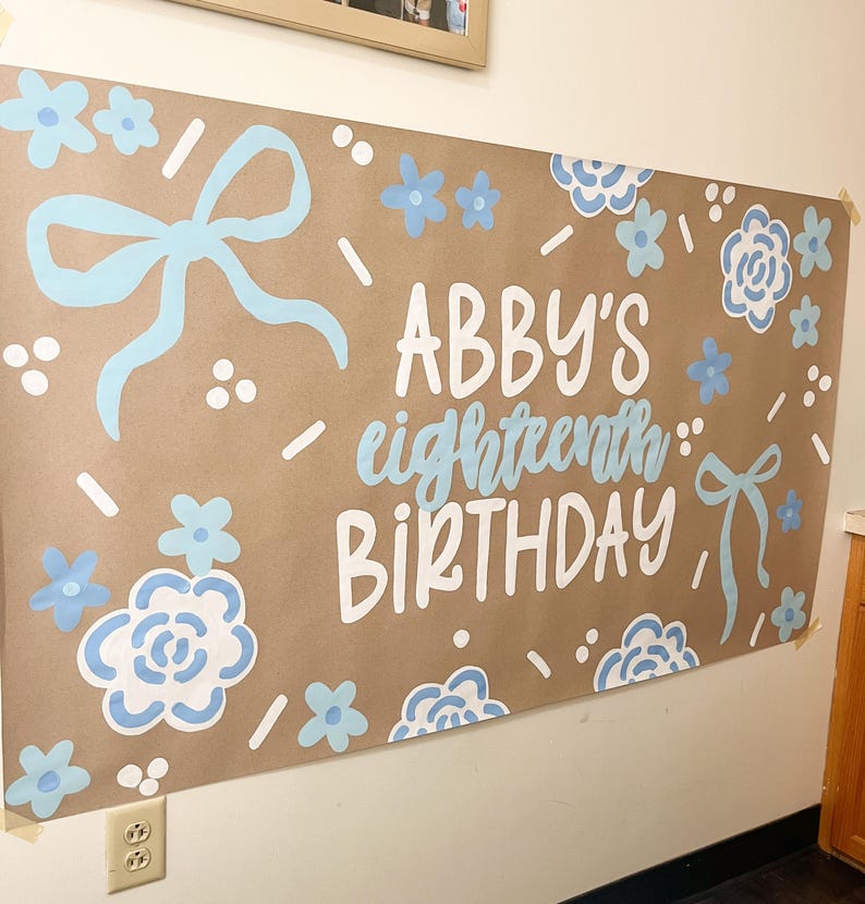 May include: A large brown paper banner with the words "ABBY'S eighteenth BIRTHDAY" in white and light blue lettering. The banner is decorated with light blue bows, flowers, and confetti-like accents. The banner is taped to a white wall.