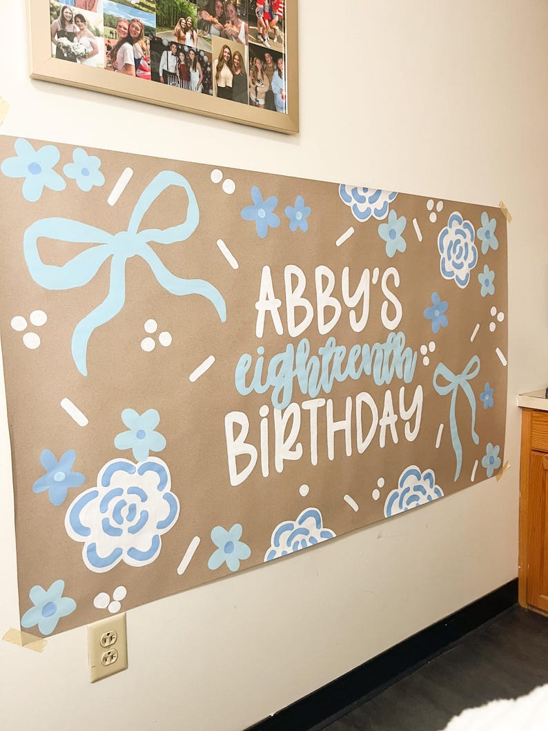 May include: A brown paper banner with the text "ABBY'S eighteenth BIRTHDAY" in white and light blue. The banner is decorated with light blue bows and floral designs. A framed photo collage hangs above.