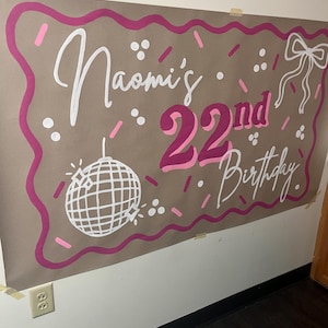 May include: A large brown paper banner with pink and white lettering and decorations. The banner reads "Naomi's 22nd Birthday" in a playful font. A disco ball and bow are also featured in the design.