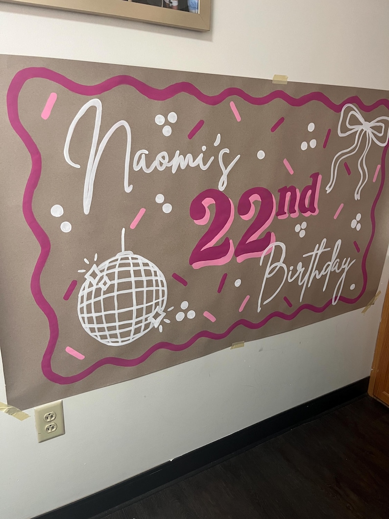 May include: A large brown paper banner with pink and white lettering and decorations. The banner reads "Naomi's 22nd Birthday" in a playful font. A disco ball and bow are also featured in the design.