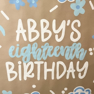 May include: A brown poster with the words "ABBY'S eighteenth BIRTHDAY" in white and light blue lettering. The poster is decorated with light blue flowers and cloud-like shapes. The overall theme is a birthday celebration.