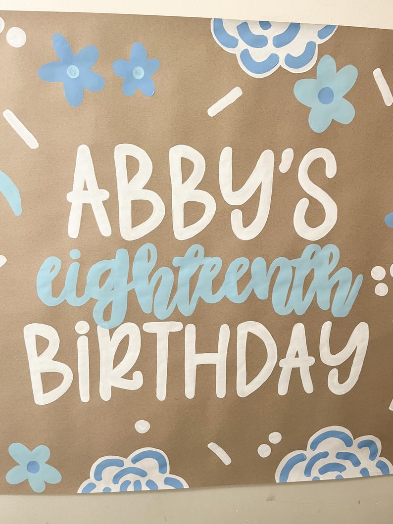 May include: A brown poster with the words "ABBY'S eighteenth BIRTHDAY" in white and light blue lettering. The poster is decorated with light blue flowers and cloud-like shapes. The overall theme is a birthday celebration.
