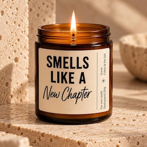 Könnte beinhalten: Eine brennende Sojawachskerze in einem bernsteinfarbenen Glasgefäß mit cremefarbenem Etikett. Das Etikett trägt den schwarzen Text "SMELLS LIKE A New Chapter" und "Scent: Cotton & Sea Salt" sowie "Hand poured | 550g". Die Kerze hat einen Docht.