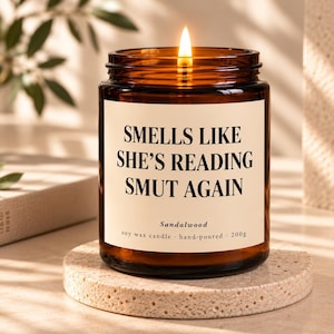 Könnte beinhalten: Eine brennende Sojawachskerze in einem bernsteinfarbenen Glasgefäß mit cremefarbenem Etikett und dem schwarzen Text: SMELLS LIKE SHE'S READING SMUT AGAIN, Sandalwood, soy wax candle, hand-poured, 200g. Die Kerze steht auf einem runden, strukturierten beigen Steinuntersetzer.